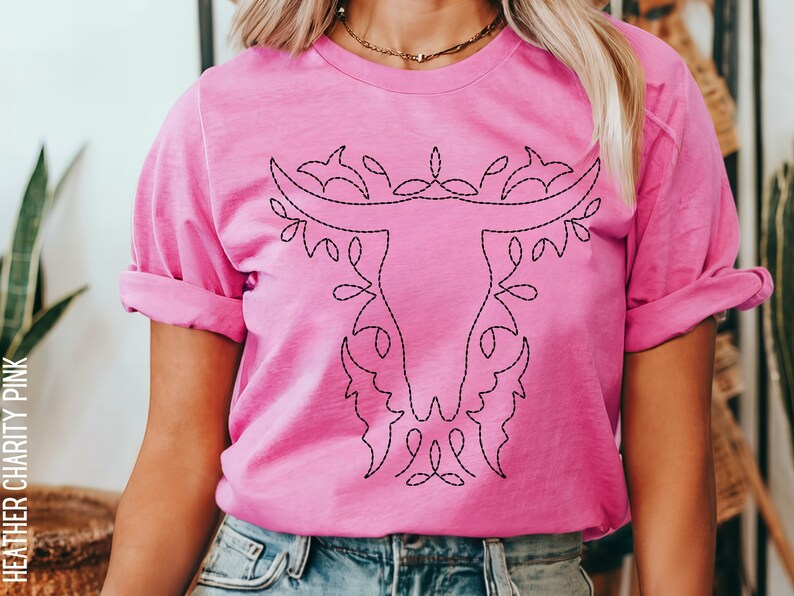 May include: A pink t-shirt with a black embroidered design of a stylized bull's head with floral accents. The text "HEATHER CHARITY PINK" is visible on the left side of the image.