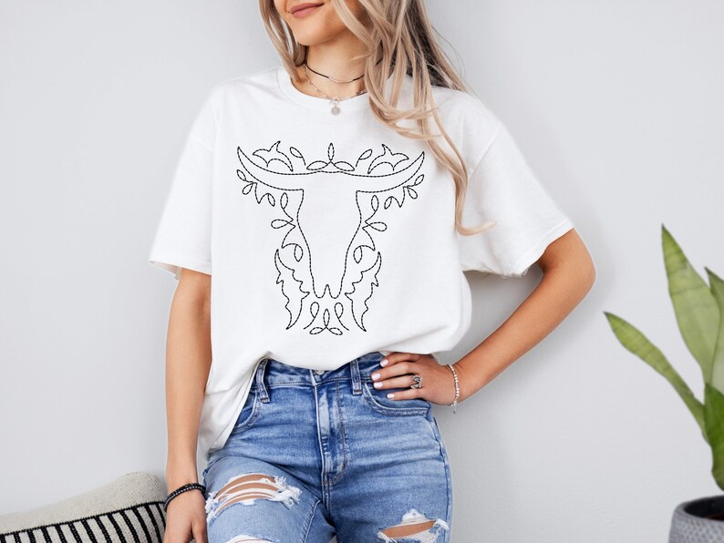May include: White t-shirt with a black embroidered design of a bull skull with floral accents.