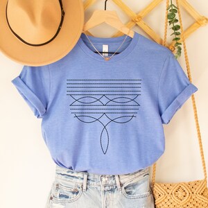 Puede incluir: Una camiseta azul cielo jaspeada con un dise&ntilde;o geom&eacute;trico en negro. El dise&ntilde;o presenta una serie de l&iacute;neas horizontales y formas curvas.