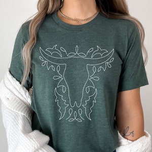 May include: A green t-shirt with a white embroidered bull's head design. The design features a stylized bull's head with floral accents.