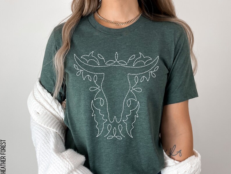 May include: A green t-shirt with a white embroidered bull's head design. The design features a stylized bull's head with floral accents.