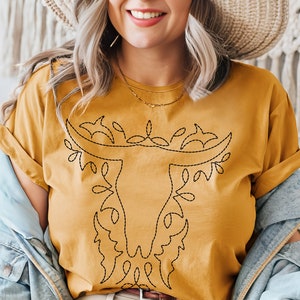 May include: A mustard yellow t-shirt with a black embroidered design of a stylized bull's head with floral accents.