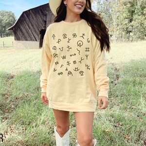 Op de afbeelding: Een geel sweatshirt met een cirkelvormig ontwerp van zwarte cowboymerken. Het sweatshirt wordt gedragen door een persoon die in een veld staat met een schuur op de achtergrond.