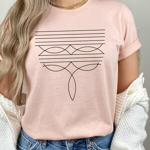 Puede incluir: Una camiseta rosa con un dise&ntilde;o geom&eacute;trico en hilo negro. El dise&ntilde;o presenta una serie de l&iacute;neas horizontales y l&iacute;neas curvas que crean un patr&oacute;n sim&eacute;trico.