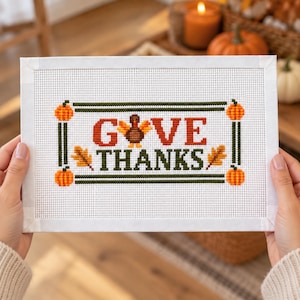 Peut inclure: Une broderie au point de croix rectangulaire sur tissu Aida blanc avec le texte GIVE THANKS en orange et vert. Le motif comprend une dinde brune remplaçant la lettre I, quatre citrouilles orange aux coins et deux feuilles d'automne dorées.