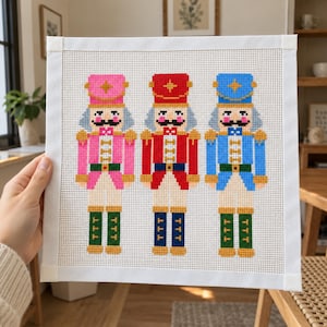 Könnte beinhalten: Ein quadratisches Stickbild mit einem Kreuzstichmuster aus drei Nussknacker-Figuren in rosa, roten und blauen Uniformen. Jeder Nussknacker hat einen Schnurrbart, einen hohen Hut mit einem Stern und grüne oder blaue Stiefel auf weißem Gittergrund.