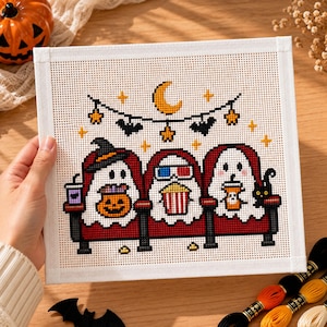May include: A square cross-stitch embroidery piece on white fabric featuring three white ghosts sitting in red theater seats. The ghosts are holding a pumpkin bucket, popcorn, and a drink. Above them is a crescent moon and a string of star and bat lights.