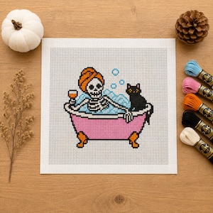 May include: A cross-stitch pattern on white fabric featuring a skeleton with an orange towel wrapped around its head, relaxing in a pink clawfoot bathtub filled with blue bubbles, holding a glass, and accompanied by a black cat.
