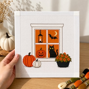 Peut inclure: Une broderie au point de croix carrée sur tissu blanc représentant une fenêtre avec un fond orange. Le motif comprend une lanterne noire, un croissant de lune, une chauve-souris, une citrouille sculptée, un chat noir, deux citrouilles et une fleur orange en pot.
