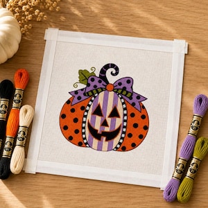 Peut inclure: Une toile de canevas carrée présentant un motif central de citrouille d'Halloween avec des pois orange, un visage rayé violet et blanc, et un nœud à pois violets. La toile est entourée de plusieurs écheveaux de fil à broder orange, crème, noir, violet et vert.