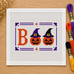 Könnte beinhalten: Ein quadratisches Stickbild mit einem Kreuzstichmotiv des Wortes BOO, bei dem die beiden Os durch orangefarbene Kürbisse mit violetten und schwarzen Hexenhüten ersetzt sind, alles umschlossen von einem violetten und orangefarbenen rechteckigen Rahmen.