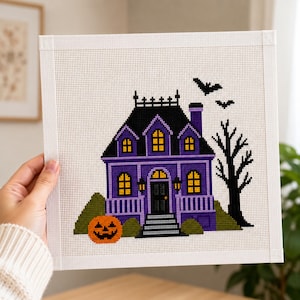 May include: A square needlepoint canvas featuring a purple haunted house with a black roof, yellow windows, and a black door. An orange jack-o'-lantern sits in the foreground, while a black leafless tree and two flying bats are on the right side.