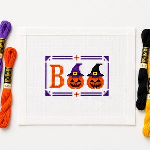 Könnte beinhalten: Ein Kreuzstichmuster auf weißem Aida-Stoff mit dem Wort BOO, bei dem die beiden Os orangefarbene Kürbisse mit violetten und schwarzen Hexenhüten sind. Das Design ist von einem violetten Rahmen umgeben und von Stickgarnsträngen flankiert.
