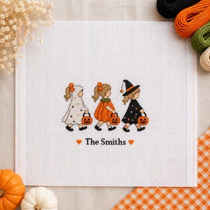 May include: A white needlepoint canvas featuring a cross-stitch design of three children in Halloween costumes: a ghost, a pumpkin, and a witch. Each child carries an orange jack-o'-lantern bucket. The text "The Smiths" is centred below.
