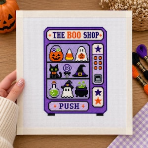 Op de afbeelding: Een kruissteekpatroon op witte stof met een paarse automaat met de tekst THE BOO SHOP en PUSH. De automaat toont Halloween-iconen waaronder een pompoen, candy corn, een spook, een zwarte kat, een lolly, een heksenhoed, een drankje en een ketel.