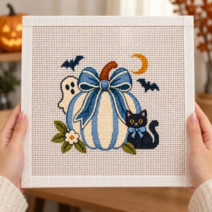 Peut inclure: Une toile de canevas carrée présentant un motif au point de croix d'une citrouille blanche à rayures bleues avec un grand nœud bleu. La scène inclut un petit fantôme blanc, un chat noir avec un nœud bleu, deux chauves-souris, un croissant de lune jaune.