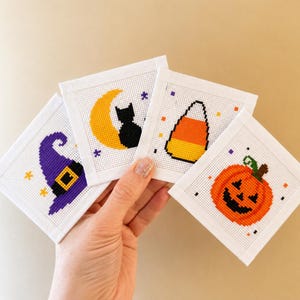 May include: Four square white fabric pieces featuring Halloween-themed cross-stitch designs. The patterns include a purple witch hat, a black cat sitting on a yellow crescent moon, a piece of candy corn, and an orange jack-o'-lantern pumpkin.