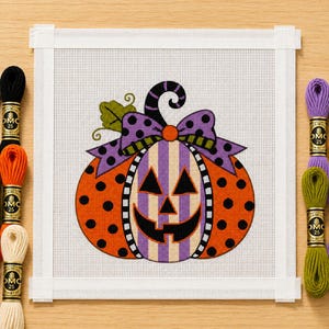 May include: A needlepoint canvas featuring a jack-o'-lantern pumpkin with orange polka dots, a purple and white striped centre, and a purple bow with black dots. The design includes a green leaf and is surrounded by six skeins of embroidery floss.
