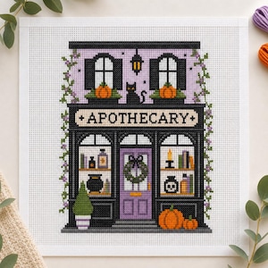 May include: A cross-stitch pattern on white Aida cloth depicting a two-storey apothecary shop. The building features a purple facade, black trim, a central door with a wreath, and windows displaying pumpkins, a black cat, and potion bottles.