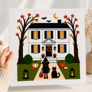 May include: A cross-stitch pattern on white fabric depicting a two-storey house with black shutters, orange glowing windows, and a front door with a wreath. A figure in a witch hat and a black cat stand on a path with pumpkins and lanterns.