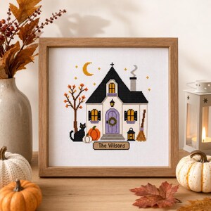 Könnte beinhalten: Ein gerahmtes Kreuzstichbild mit einem weißen Haus mit schwarzem Dach, einer lila Tür, einer schwarzen Katze, Kürbissen und einem Besen. Das Design enthält einen gelben Halbmond, Sterne und ein Schild mit der Aufschrift The Wilsons.