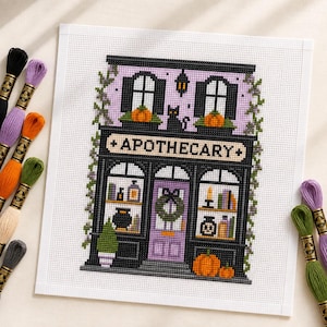 Peut inclure: Un motif de point de croix sur toile Aida blanche représentant une boutique d'apothicaire à deux étages avec une devanture noire, une porte violette avec une couronne, des citrouilles orange et un chat noir. Le mot APOTHECARY est brodé en noir. Des échevettes entourent le motif.