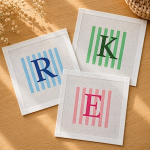 May include: Three square needlepoint canvases featuring embroidered letters R, K, and E. Each letter is centred over a background of vertical stripes in blue, green, and pink respectively. The white mesh fabric has a distinct grid texture.