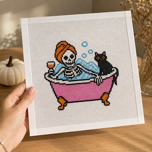 May include: A square needlepoint canvas featuring a cross-stitch design of a skeleton wearing an orange towel wrap on its head, relaxing in a pink clawfoot bathtub filled with blue water and bubbles, holding a glass, with a black cat sitting nearby.