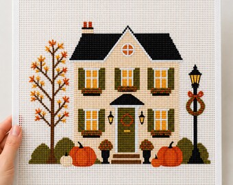 May include: A cross-stitch pattern on white fabric featuring a two-story house with a black roof, beige walls, and green shutters. The scene includes a tall tree with orange leaves, a black lamppost with a wreath, and several orange pumpkins.