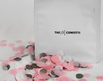 Puede incluir: Una bolsa blanca con el texto "THE KM CONFETTI" impreso en ella. La bolsa está abierta y se derrama confeti rosa, blanco y gris.