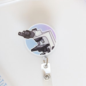 May include: A round, white and purple microscope badge reel with a clear plastic strap and metal clip. The badge reel features a cartoon illustration of a microscope. Other lab equipment is visible in the background.