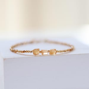 May include: A delicate gold bracelet featuring small, round beads and three larger, rectangular amber-colored gemstones. The bracelet is displayed on a white surface, with a soft, blurred background.