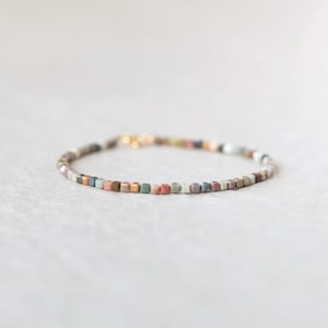 May include: A delicate bracelet featuring small, square beads in a variety of muted colors, including pink, green, and brown. The bracelet has a gold-colored clasp. The beads are approximately 0.12 inches square.