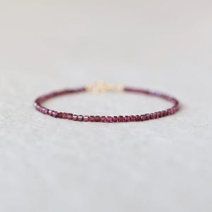 May include: A delicate bracelet featuring small, square, deep red beads strung together. The bracelet has a gold-colored clasp and is set against a soft, neutral background. The beads appear to be approximately 0.1 inches in size.