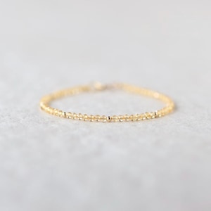May include: Delicate gold bracelet featuring small, round, yellow beads. The bracelet has a simple design with a thin chain and is likely made of gold-toned metal. The beads are evenly spaced, creating a minimalist and elegant look.