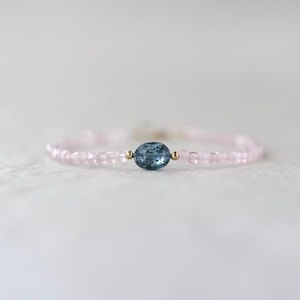 May include: Delicate pink beaded bracelet with a central oval blue gemstone and small gold accents. The bracelet is strung on a thin, flexible cord, and the beads are evenly spaced. The overall design is simple and elegant.