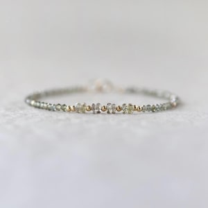 May include: Delicate bracelet featuring small, faceted, pale green gemstones interspersed with gold-colored beads. The bracelet is strung on a thin, silver-colored chain, creating a minimalist and elegant design. The bracelet is suitable for everyday wear.