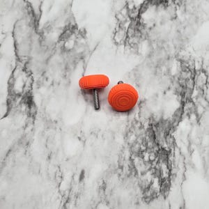 Könnte beinhalten: Zwei orangefarbene Schrankknöpfe mit kreisförmigem Design. Ein Knopf ist an einer silbernen Schraube befestigt. Der andere Knopf hat eine runde Form. Die Knöpfe befinden sich auf einer weiß-grauen Marmoroberfläche.