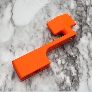 May include: Orange plastic safety device for a firearm. The device has a lever and a small handle.