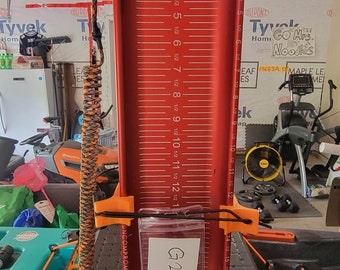 May include: A red measuring stick with a white label that says "G24" and a black and orange rope attached to it. The measuring stick has a white label that says "Rebel RB Bourbon" and a white label that says "Ketch". The measuring stick is attached to a black and orange kayak.