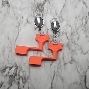 May include: Two orange keychains with a retractable cord and a metal clip. The keychains have a unique T-shaped design with a textured grip. The background is a marble pattern.