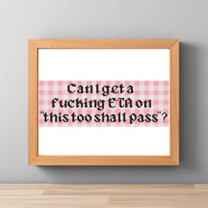 Könnte beinhalten: Eine gerahmte Kreuzstichstickerei mit einem rosa-weiß karierten Hintergrundmuster und schwarzem, verpixeltem Text, der lautet: Can't get a fucking ETA on "this too shall pass"? Das Kunstwerk befindet sich in einem hellen Holzrahmen.