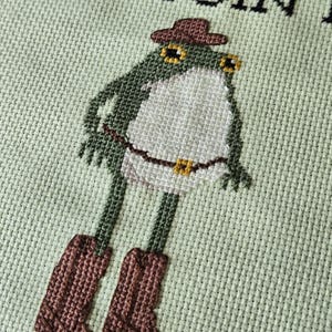 May include: Cross-stitch design of a frog wearing a cowboy hat and boots. The frog is green and white, with brown boots and a brown hat. The background is a light green fabric with the text "ONLY SO I'M DOIN".