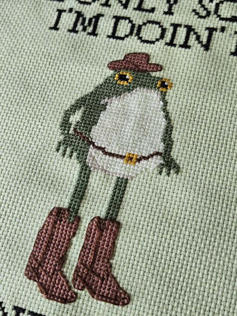 May include: Cross-stitch design of a frog wearing a cowboy hat and boots. The frog is green and white, with brown boots and a brown hat. The background is a light green fabric with the text "ONLY SO I'M DOIN".