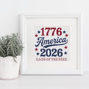 May include: A framed cross-stitch artwork with the text "1776 America 2026 Land of the Free" in red and blue, surrounded by stars. The artwork is in a white frame, and a potted plant is visible on the left.