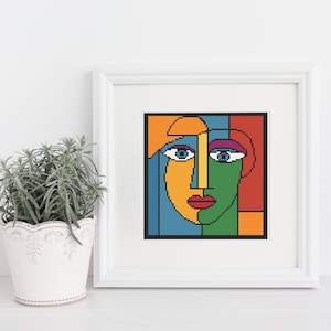May include: A framed abstract cross-stitch art piece featuring a colorful face. The face is divided into sections of orange, yellow, green, blue, and red. The eyes are blue with white highlights, and the lips are red. The artwork is in a white frame.
