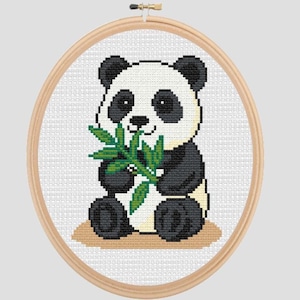 May include: Cross-stitch embroidery of a panda bear holding green bamboo leaves. The panda is black and white with a tan base, set in a wooden embroidery hoop. The background is white.