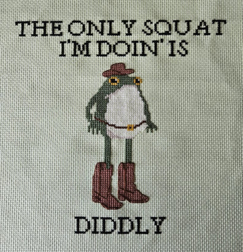 May include: Cross-stitch art featuring a green frog wearing a cowboy hat and boots, with the text "THE ONLY SQUAT I'M DOIN' IS DIDDLY" in black. The frog has a white belly and a brown belt. The background is a light green fabric.