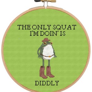 May include: Cross-stitch embroidery hoop featuring a frog wearing a cowboy hat and boots. The text reads "THE ONLY SQUAT I'M DOIN' IS DIDDLY" in black letters on a green background. The hoop is made of wood.