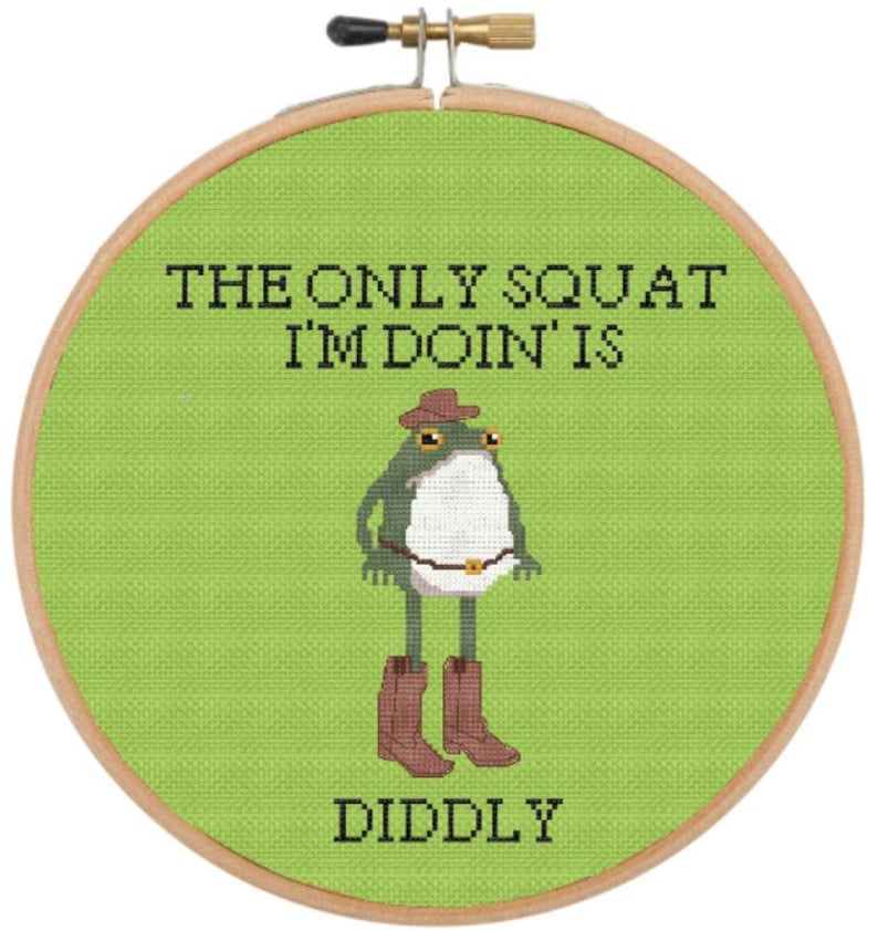 May include: Cross-stitch embroidery hoop featuring a frog wearing a cowboy hat and boots. The text reads "THE ONLY SQUAT I'M DOIN' IS DIDDLY" in black letters on a green background. The hoop is made of wood.